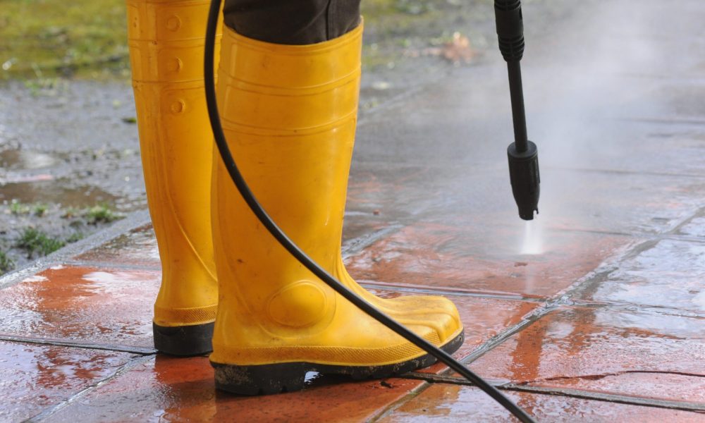 A person wearing yellow rubber boots with high-pressure water nozzle cleaning the dirt in the tiles